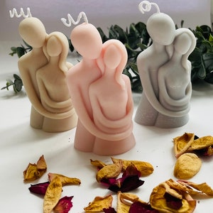 Puede incluir: Tres velas de pareja abraz&aacute;ndose en beige, rosa y gris. Cada vela presenta una figura masculina y femenina abraz&aacute;ndose. Las velas tienen mechas blancas y rizadas. P&eacute;talos de rosa secos esparcidos alrededor de las velas.