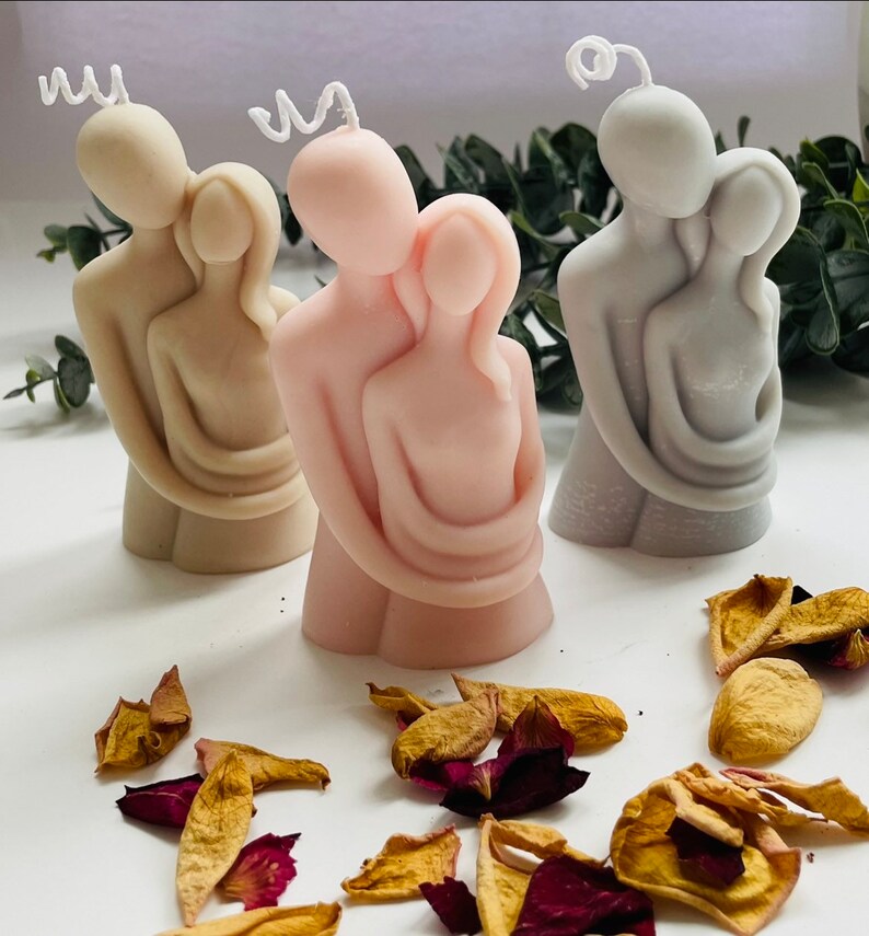 Puede incluir: Tres velas de pareja abraz&aacute;ndose en beige, rosa y gris. Cada vela presenta una figura masculina y femenina abraz&aacute;ndose. Las velas tienen mechas blancas y rizadas. P&eacute;talos de rosa secos esparcidos alrededor de las velas.