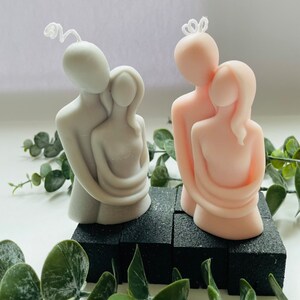 Puede incluir: Dos velas decorativas con forma de figuras abrazadas. Una vela es gris claro, la otra rosa claro. Ambas velas tienen una mecha blanca y est&aacute;n sobre una base negra. La vegetaci&oacute;n rodea las velas.