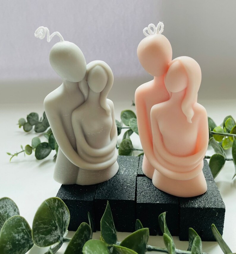 Puede incluir: Dos velas decorativas con forma de figuras abrazadas. Una vela es gris claro, la otra rosa claro. Ambas velas tienen una mecha blanca y est&aacute;n sobre una base negra. La vegetaci&oacute;n rodea las velas.