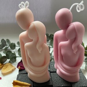 Puede incluir: Dos velas rosas y rosa claro que representan figuras abrazadas. Cada vela tiene una mecha blanca y se asienta sobre una base negra texturizada. Las velas est&aacute;n rodeadas de vegetaci&oacute;n y p&eacute;talos de flores secas.