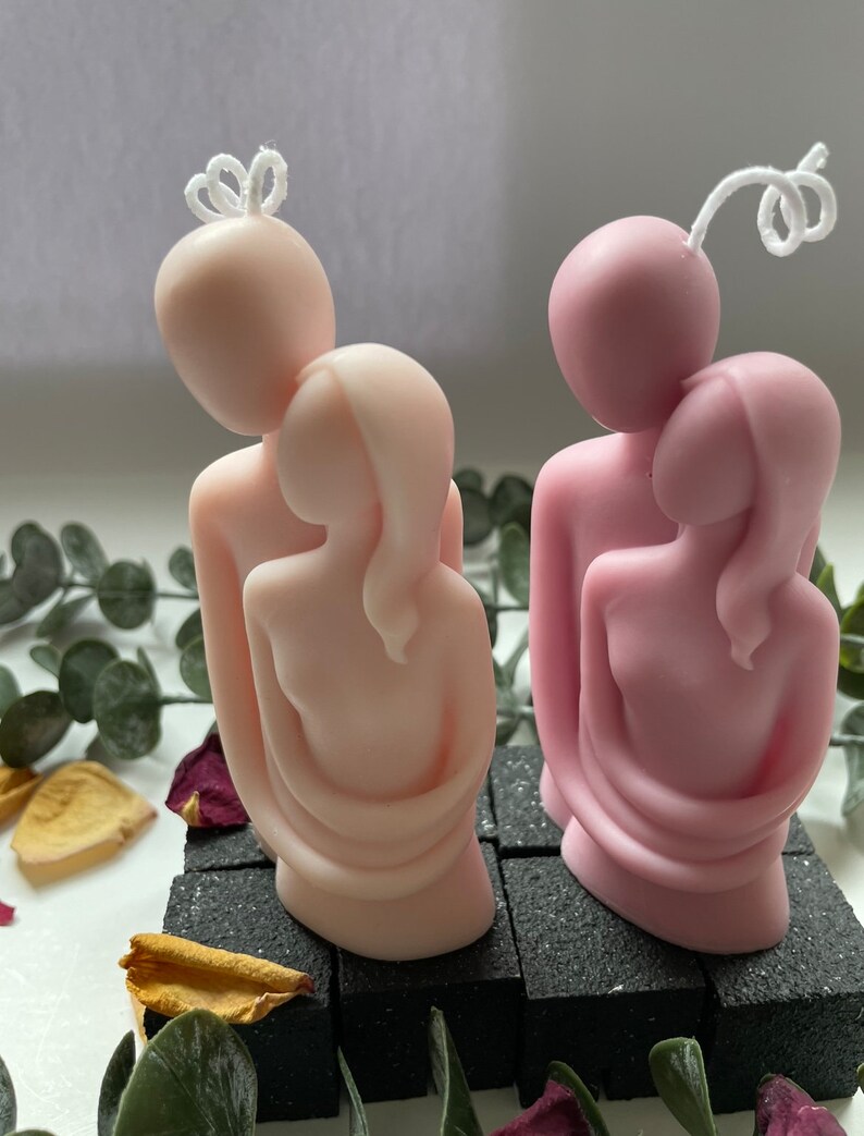 Puede incluir: Dos velas rosas y rosa claro que representan figuras abrazadas. Cada vela tiene una mecha blanca y se asienta sobre una base negra texturizada. Las velas est&aacute;n rodeadas de vegetaci&oacute;n y p&eacute;talos de flores secas.
