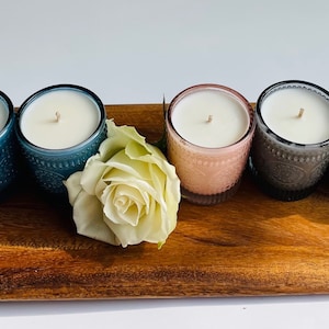 Puede incluir: Un conjunto de siete velas de varios colores, incluyendo azul verdoso, gris y rosa, dispuestas en una bandeja de madera. Las velas están en frascos de vidrio con patrones decorativos.