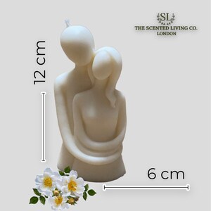 Puede incluir: Vela de color marfil que representa una pareja abraz&aacute;ndose, mide 12 cm de alto y 6 cm de ancho. La vela tiene un acabado liso y est&aacute; acompa&ntilde;ada de un arreglo floral. Se ve el logo de The Scented Living Co. London.