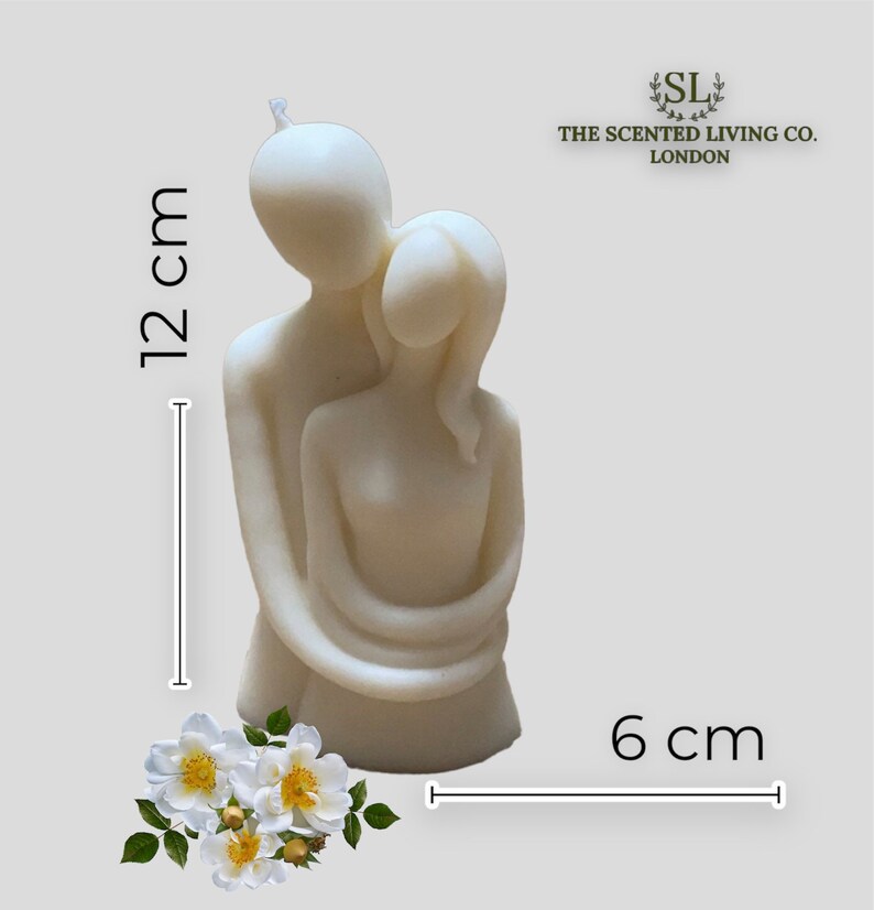 Puede incluir: Vela de color marfil que representa una pareja abraz&aacute;ndose, mide 12 cm de alto y 6 cm de ancho. La vela tiene un acabado liso y est&aacute; acompa&ntilde;ada de un arreglo floral. Se ve el logo de The Scented Living Co. London.