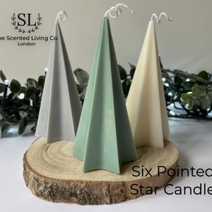Puede incluir: Tres velas en forma de estrella de seis puntas en gris, verde y blanco. Las velas están dispuestas sobre una rodaja de madera. El texto "Six Pointed Star Candle" está impreso debajo de las velas.