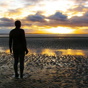Könnte beinhalten: Eine Silhouette einer Person, die auf einem Sandstrand steht und zum Meer blickt. Die Sonne geht hinter der Person unter und wirft einen goldenen Schein auf das Wasser und den Sand. Der Himmel ist eine Mischung aus Orange, Gelb und Blau.
