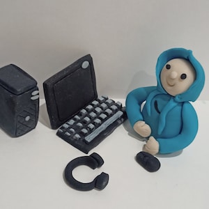 May include: A blue figurine of a person wearing a hoodie, sitting at a computer with a black desktop computer, a black laptop, a black keyboard, and a black headset.