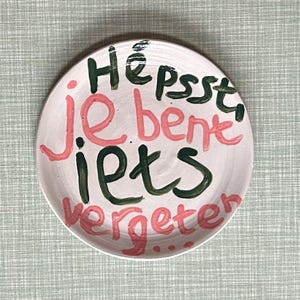 Könnte beinhalten: Ein runder, cremefarbener Keramikteller mit den Worten "Hè psst, je bent iets vergeten..." in rosa und grüner Handschrift. Der Teller hat eine einfache, handgemachte Ästhetik.