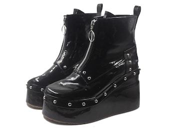 May include: A pair of black patent leather ankle boots featuring a high platform sole, a front-facing silver zipper closure with a circular pull tab, and silver-toned metal studs lining the base and side panels of the footwear.