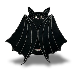 May include: A black bat-shaped bag featuring large, spread wings with white stitched detailing. The face has two silver metal snap buttons for eyes, a small pink heart-shaped nose, and two pointed ears. A small, colourful logo is visible at the base.