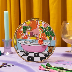 May include: A circular embroidery hoop showing a bathroom scene with a pink bathtub, plants, and a wine glass. The background has an orange and red design. A green candle and a gold glass are also in the image.