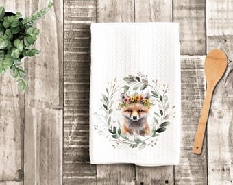May include: White kitchen towel with a textured pattern. The towel features a watercolor illustration of a fox wearing a floral crown, surrounded by a wreath of green leaves. A wooden spoon rests on a wooden surface.