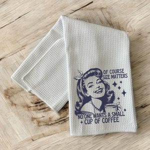 May include: A white kitchen towel with a blue retro-style illustration of a woman with a large cup of coffee. The text reads "Of course size matters. No one wants a small cup of coffee."