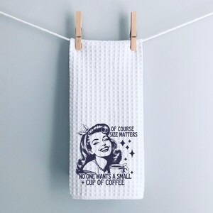 May include: White waffle weave kitchen towel with a black and white illustration of a woman holding a coffee cup. The text reads "Of course size matters. No one wants a small cup of coffee."