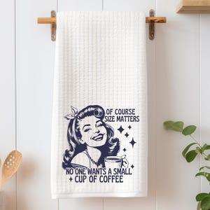 May include: White kitchen towel with a black and white illustration of a woman holding a coffee mug. The text reads "Of course size matters. No one wants a small cup of coffee."