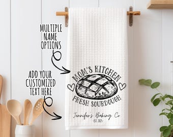 May include: White kitchen towel with black text that reads "Mom's Kitchen Fresh Sourdough" with a drawing of a loaf of bread and a heart. The text also includes "Jennifer's Baking Co. -Est. 2025-."