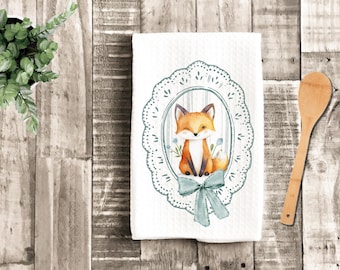 May include: White kitchen towel with a winking fox illustration inside a decorative oval frame. The fox is orange and white with a blue bow at the bottom of the frame. The towel has a textured pattern.