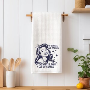 May include: White kitchen towel with a blue graphic of a woman holding a cup of coffee. The text reads "Of course size matters. No one wants a small cup of coffee."