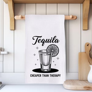May include: White kitchen towel with a black graphic of a tequila shot, a lime slice, and the words "Tequila" and "Cheaper Than Therapy". The towel is hanging on a wooden rod.