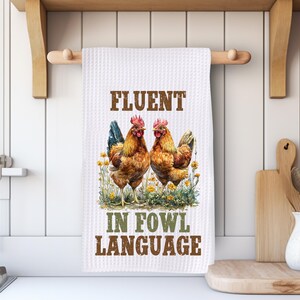 May include: White kitchen towel with the text "FLUENT IN FOWL LANGUAGE" and a watercolor illustration of two brown and red roosters standing in a field of yellow flowers. The towel is hanging on a wooden rod.