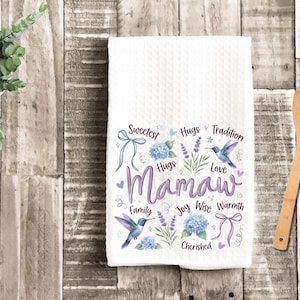 May include: White kitchen towel with a textured pattern. The towel features the word "Mamaw" in purple script, surrounded by floral and hummingbird illustrations, and words like "Sweetest" and "Love".
