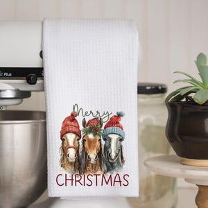 May include: White kitchen towel with a waffle weave texture. The towel features a watercolor illustration of three horses wearing red and blue winter hats. The words "Merry Christmas" are printed in red.