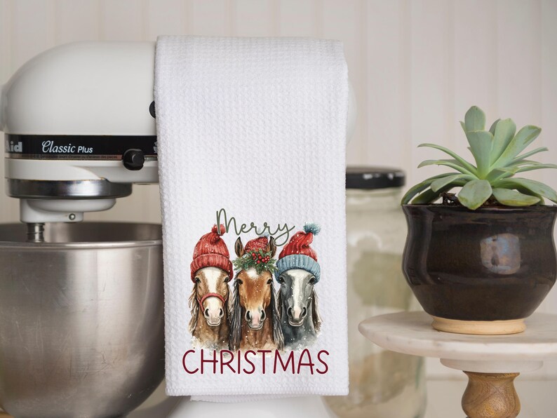 May include: White kitchen towel with a waffle weave texture. The towel features a watercolor illustration of three horses wearing red and blue winter hats. The words "Merry Christmas" are printed in red.