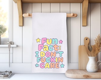 May include: White waffle-weave kitchen towel with the text "Fun Fact: I Don't Fuckin' Care" in pastel colors. The towel is hanging on a wooden rod, with a wooden shelf above and a cutting board and vase in the background.