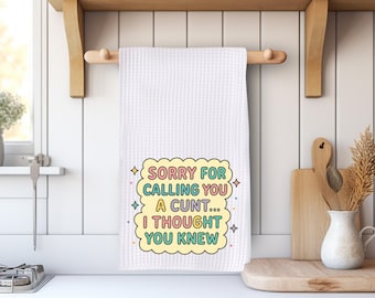 May include: White waffle-weave kitchen towel with the text "SORRY FOR CALLING YOU A CUNT... I THOUGHT YOU KNEW" in a colorful, cartoonish design. The towel is hanging on a wooden rod in a kitchen setting.