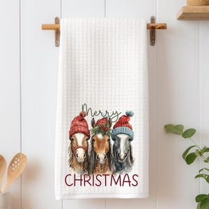 May include: White waffle-weave kitchen towel featuring a watercolor illustration of three horses wearing red and blue Christmas hats. The words "Merry Christmas" are printed in red below the horses.