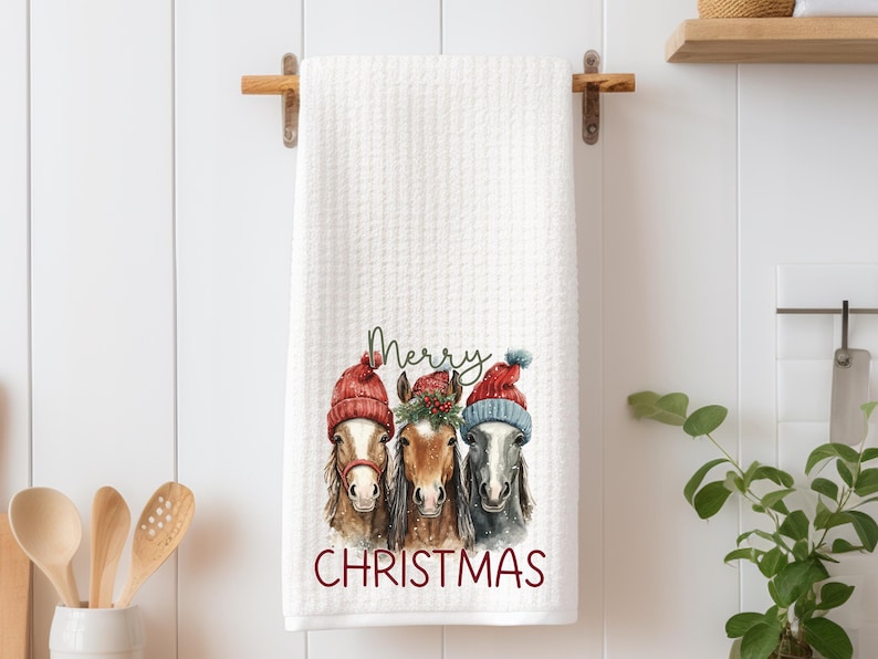 May include: White waffle-weave kitchen towel featuring a watercolor illustration of three horses wearing red and blue Christmas hats. The words "Merry Christmas" are printed in red below the horses.