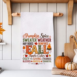 May include: A white waffle-weave kitchen towel featuring colorful autumn-themed text and graphics including the words Pumpkin Spice, Sweater Weather, Hay Rides, Bonfires, Fall, Apple Cider, Cozy, Giving, Thanks, Blessed, Grateful, and Thankful.