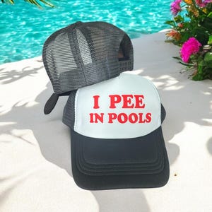 May include: A white and black trucker hat with the text "I PEE IN POOLS" in red letters. The hat is sitting on a white sandy surface.