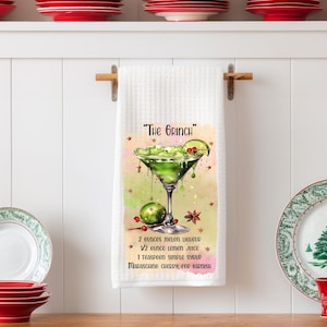 May include: White kitchen towel with a green cocktail recipe. The towel features a green cocktail illustration with the text "The Grinch" and a recipe for the cocktail. Red and green plates are in the background.