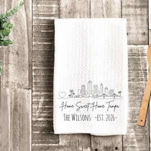 May include: A white waffle-weave kitchen towel featuring a black line art illustration of the Tampa skyline, a heart, and the text "Home Sweet Home Tampa", "THE WILSONS", and "EST. 2026" printed in a clean, dark font on the lower portion of the fabric.