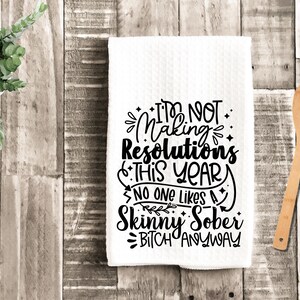 May include: White kitchen towel with black text that reads "I'm Not Making Resolutions This Year. No One Likes A Skinny Sober Bitch Anyway." A wooden spoon rests on a wooden surface.