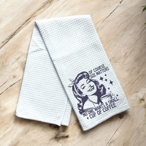 May include: White kitchen towel with a blue graphic of a woman holding a cup of coffee. The text reads "Of course size matters. No one wants a small cup of coffee."