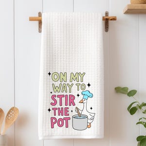 May include: White kitchen towel with a cartoon goose wearing a chef's hat and stirring a pot. The text on the towel reads "On my way to stir the pot."