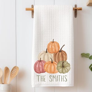 May include: White waffle-weave kitchen towel with a watercolor illustration of six pumpkins in shades of orange, pink, brown, and green. The text "THE SMITHS" is printed at the bottom. The towel is hanging on a wooden rack.