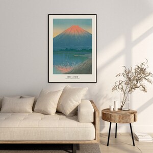 May include: Framed art print featuring a Japanese landscape with Mount Fuji. The artwork depicts the mountain with a red-orange peak, reflected in the blue water below. The print has a black frame and is displayed above a beige sofa.