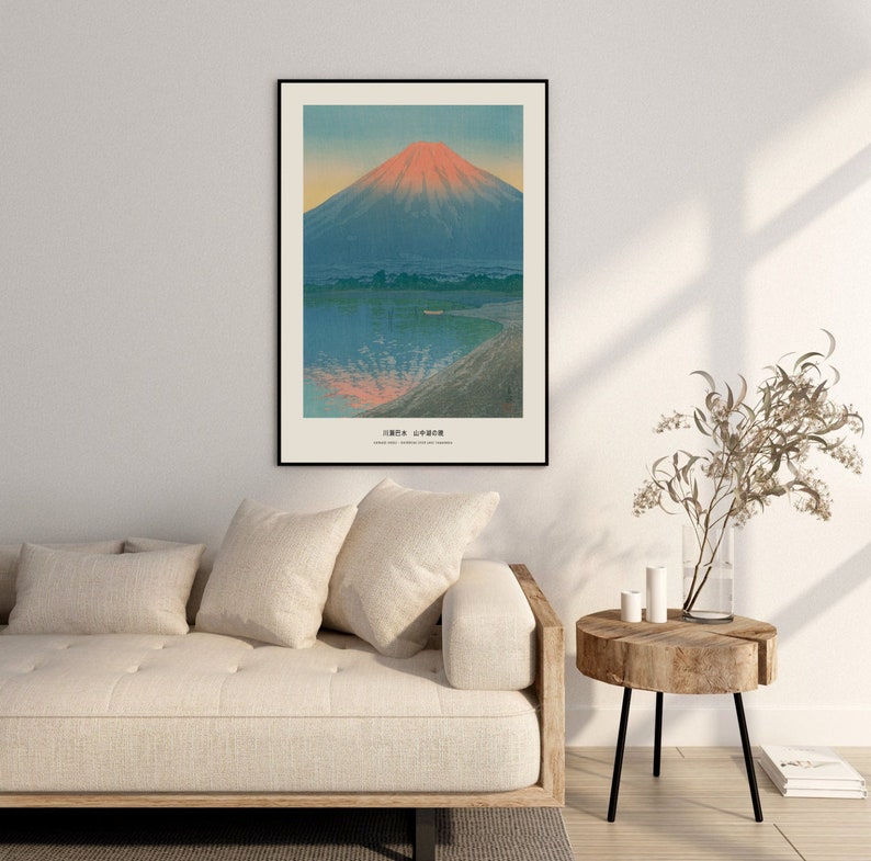 May include: Framed art print featuring a Japanese landscape with Mount Fuji. The artwork depicts the mountain with a red-orange peak, reflected in the blue water below. The print has a black frame and is displayed above a beige sofa.