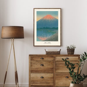 May include: A framed print of a Japanese landscape featuring Mount Fuji. The artwork depicts the mountain with a red-orange peak, set against a blue sky and water. The print is in a wooden frame and hangs above a wooden dresser.