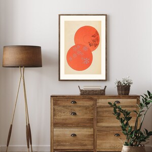 May include: A framed print of a minimalist abstract art piece featuring two overlapping circles in shades of orange and white with a pattern of small white dots. The print is displayed on a wall above a wooden dresser.