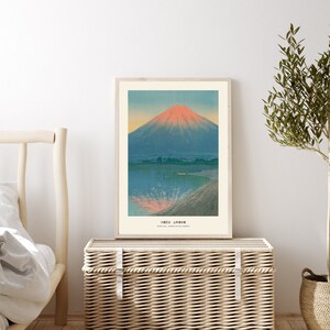May include: A framed art print featuring a mountain landscape with a lake. The mountain has a red peak, and the water reflects the colors of the sky. The print is in a light wood frame and is displayed on a woven storage chest.