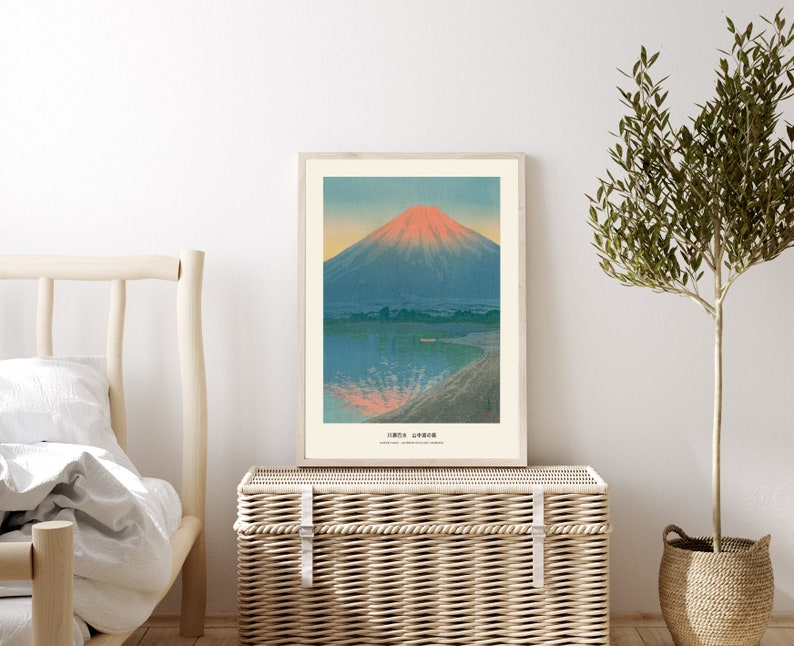May include: A framed art print featuring a mountain landscape with a lake. The mountain has a red peak, and the water reflects the colors of the sky. The print is in a light wood frame and is displayed on a woven storage chest.
