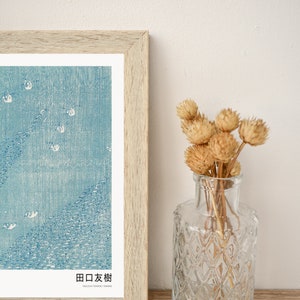 May include: A framed print of a Japanese woodblock print depicting a blue sky with white birds flying over a blue sea. The print is titled "田口友樹" in Japanese and "Yaguchi Tomoki / Shimane" in English. The print is displayed on a wooden surface next to a clear glass vase with dried flowers.