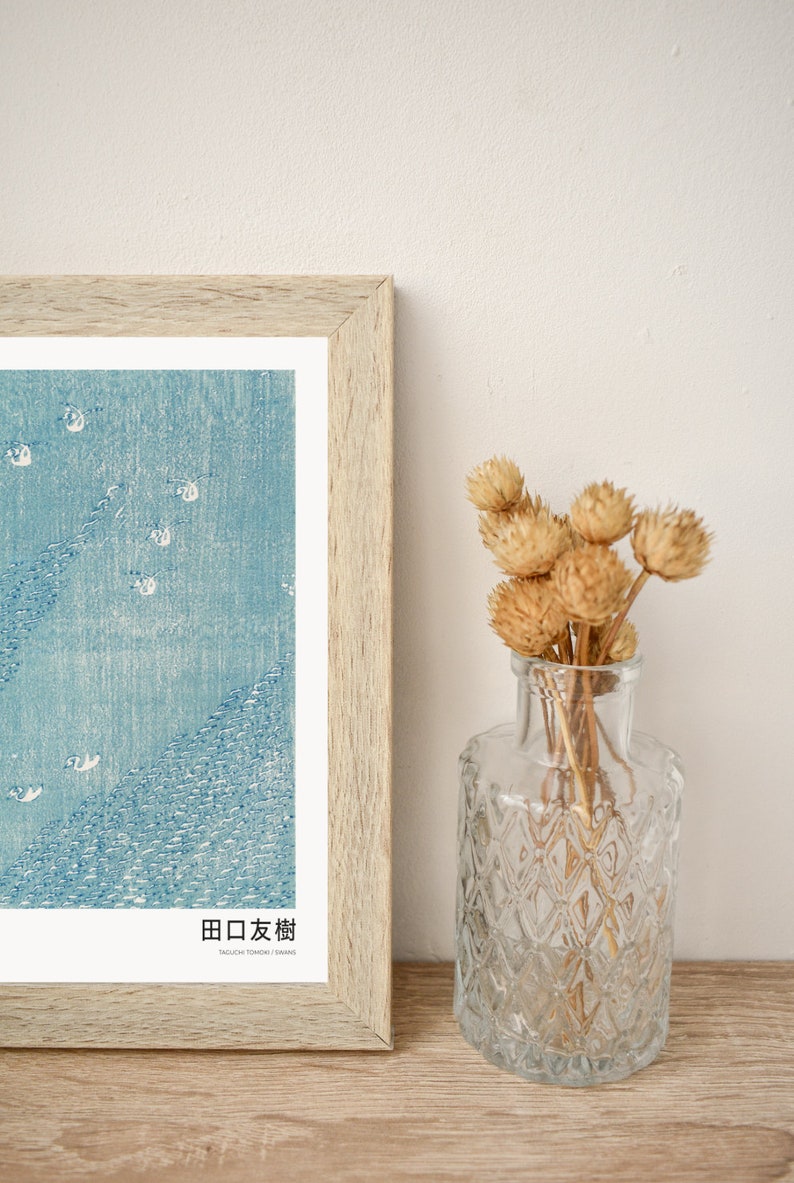 May include: A framed print of a Japanese woodblock print depicting a blue sky with white birds flying over a blue sea. The print is titled "田口友樹" in Japanese and "Yaguchi Tomoki / Shimane" in English. The print is displayed on a wooden surface next to a clear glass vase with dried flowers.