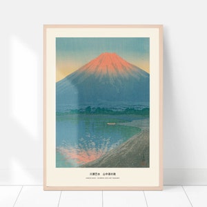 May include: A framed print of a Japanese landscape featuring Mount Fuji. The artwork depicts the mountain with a red-orange peak, reflected in the blue water. The print includes Japanese text at the bottom and is set in a light wood frame.
