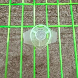 Puede incluir: Un dispositivo translúcido de color verde claro con un centro circular similar a una malla y un diseño en forma de ala. El dispositivo se encuentra sobre un fondo de un patrón de rejilla verde, lo que sugiere un tema deportivo o de equipamiento.